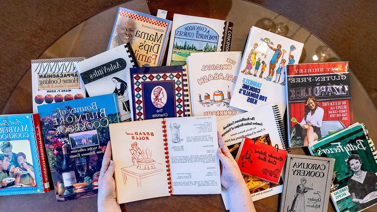 Hands holding open a vintage cookbook with a number of other cookbooks in the background.